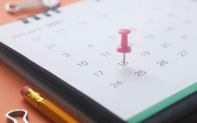 A close-up of a calendar page with a pink push pin, pencil, and binder clip on an orange background.